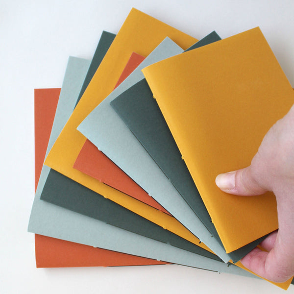 A selection of A5 and A6 exercise books in various colors including mustard, rust, lichen, and sequoia, held in a fan-like display.