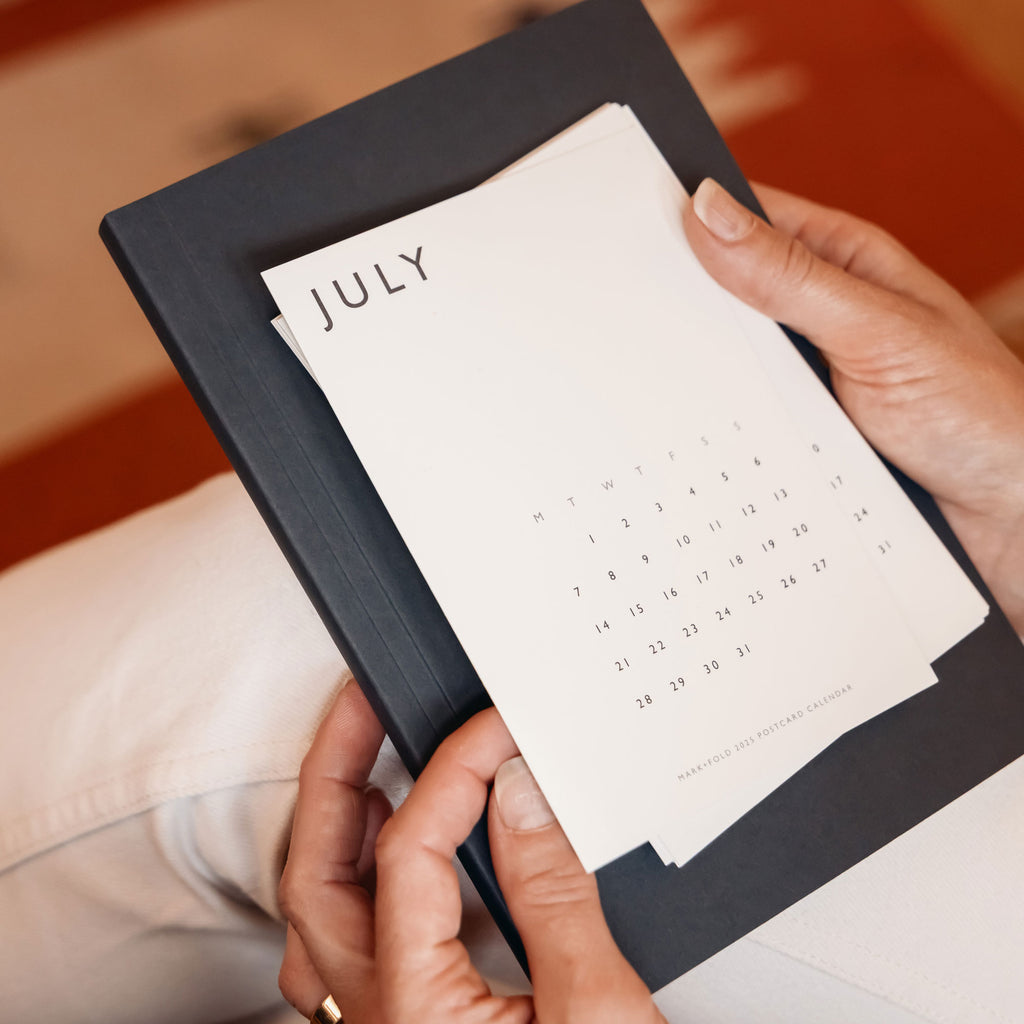 Person holding a July calendar postcard, in front of a flint notebook
