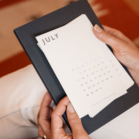Person holding a July calendar postcard, in front of a flint notebook