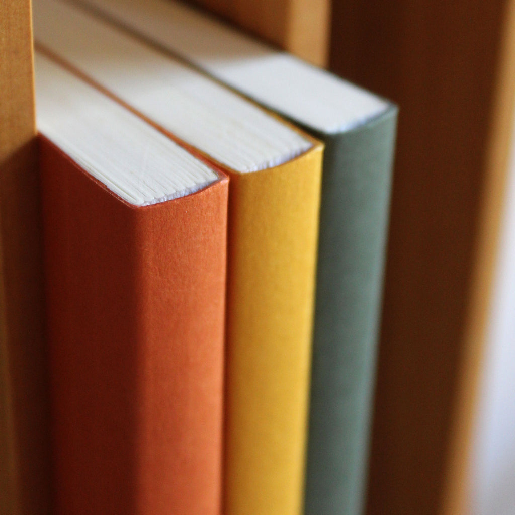 Three notebooks in rust red, mustard yellow and sequoia green, stacked on a wooden surface