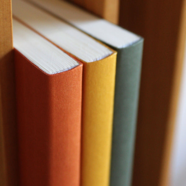 Three notebooks in rust red, mustard yellow and sequoia green, stacked on a wooden surface