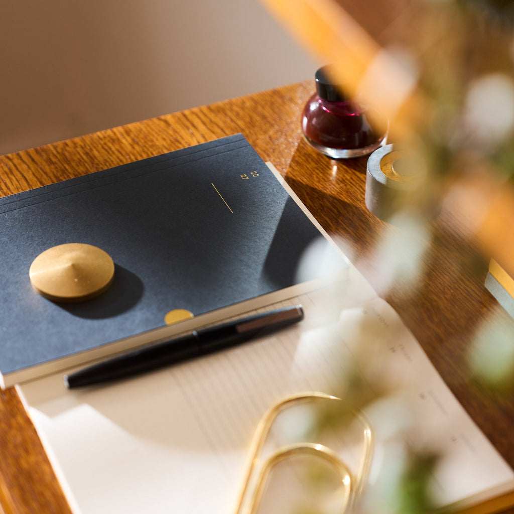 A desk scene with a navy 2027 diary, white week-view page, black pen, gold paperclip, and a small round gold object on a wooden table, softly lit with blurred foreground foliage.