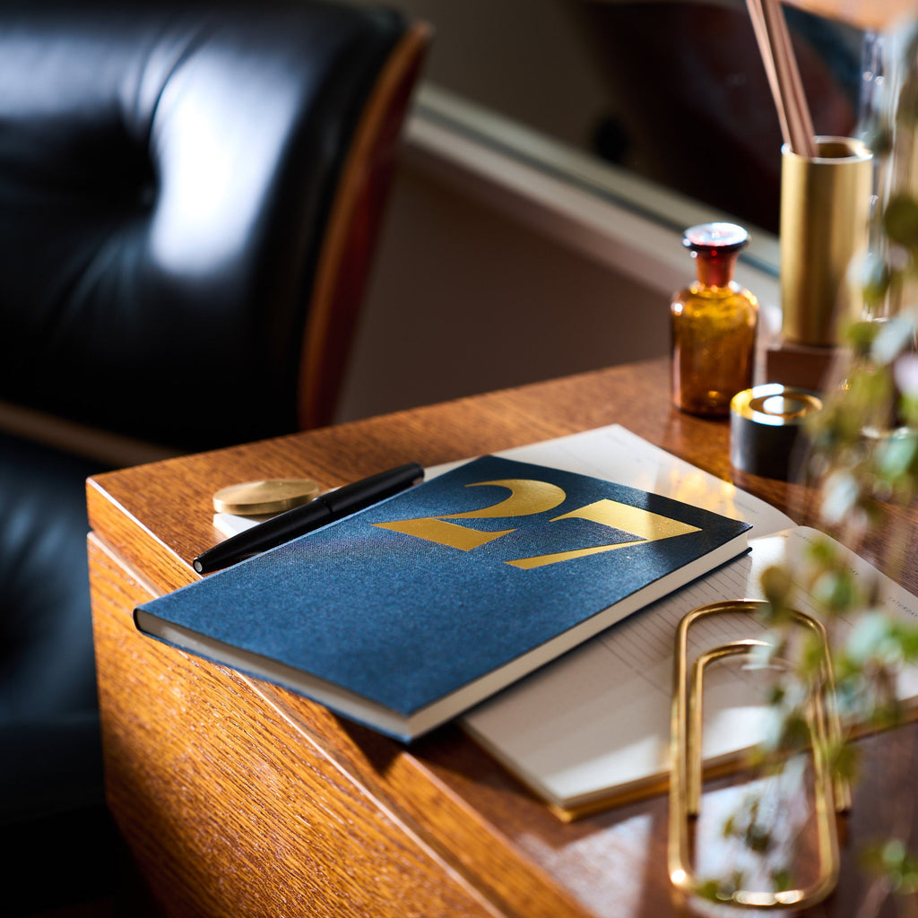 A navy blue linen planner with gold foil lettering reading '27' sits on a wooden desk next to a black leather chair, with gold and amber accessories in the background.
