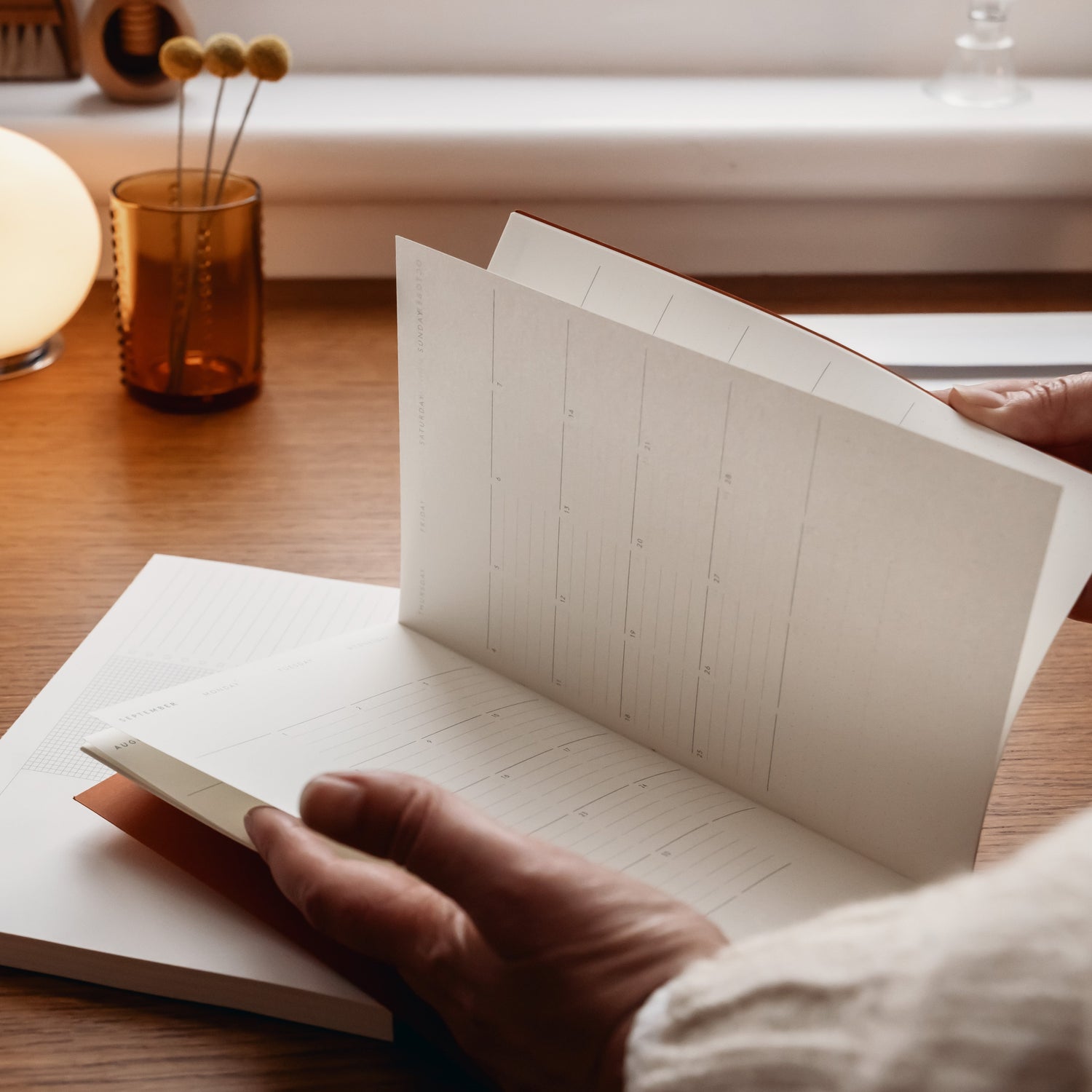 Person flicking through Monthly planner, at wooden desk with warm autumnal light and brown glass vase with dried flowers.