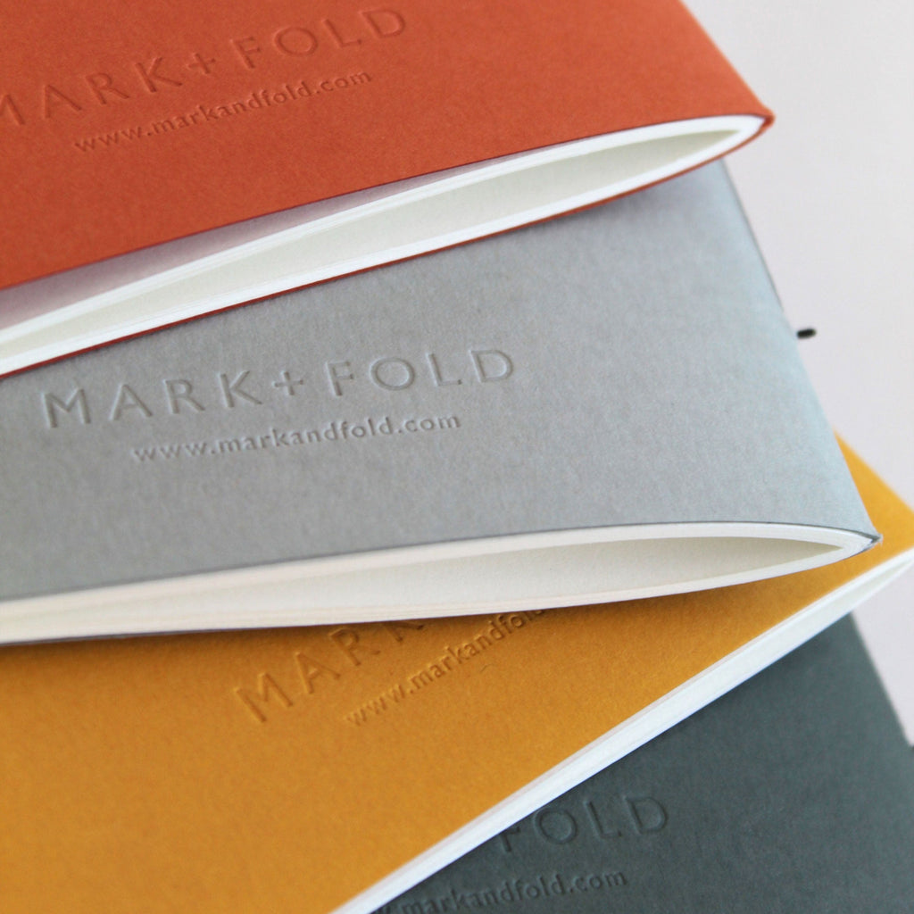 Three notebooks in orange, gray, and green with 'MARK+FOLD' blind debossed branding.