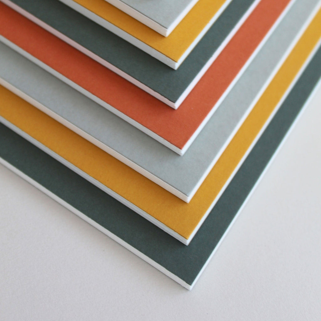 Stack of colorful slim notebooks with orange, yellow, gray, and red hues on a light gray background.