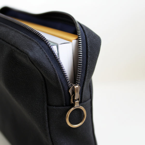 Close-up of a black canvas pouch with a brass zipper and notebooks inside, on a white background
