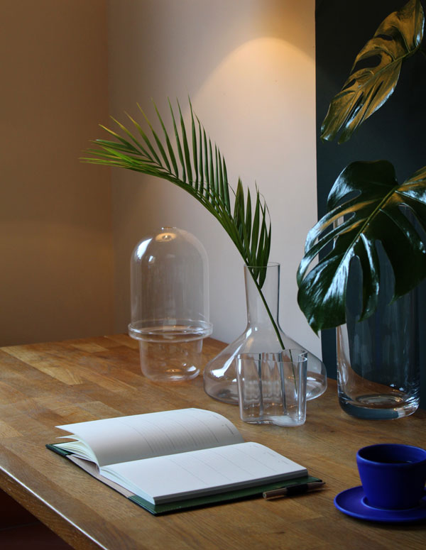 Desk scene featuring avocado green leather diary cover, blue coffee cup, glass vases and house plants